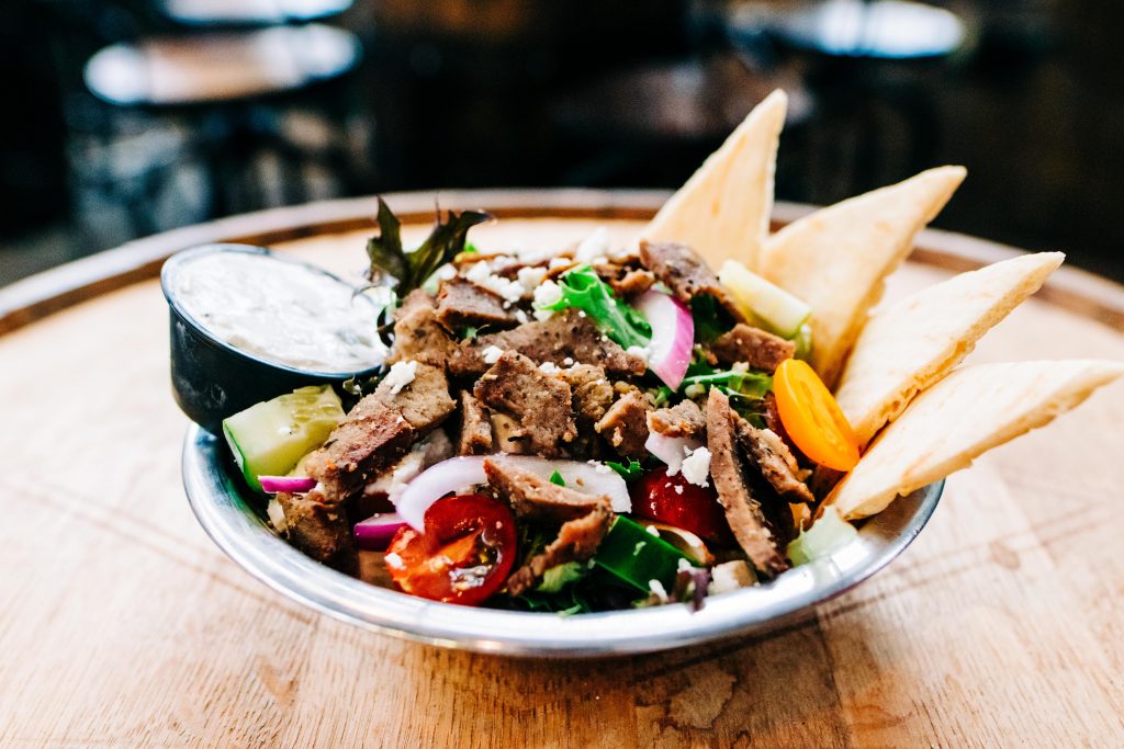 a picture of a Greek gyro salad topped with lamb meat, cucumber, onion, and tomato with tzatziki sauce and pita bread on the side